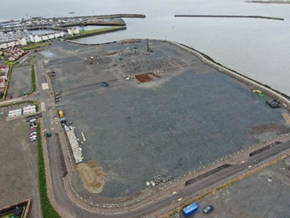 Bird's eye view of foundations of a mostly clear building site surrounded by water on one side and housing on the other.