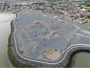 Foundations of a building site surrounded by water on one side and housing on the other.