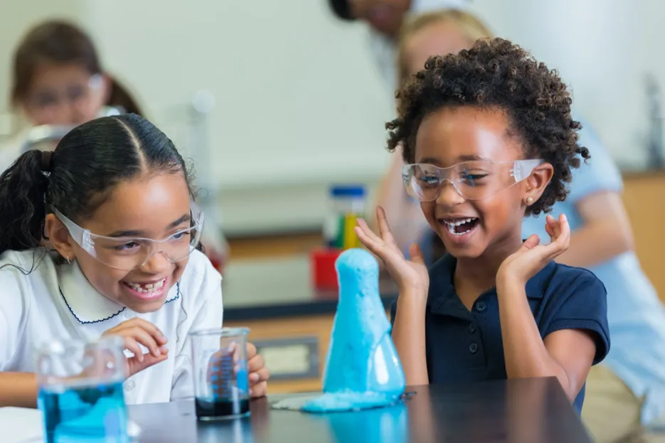 Two children laughing and looking at a chemistry experiment in a flask, there are two beakers on the table and three other people in the background