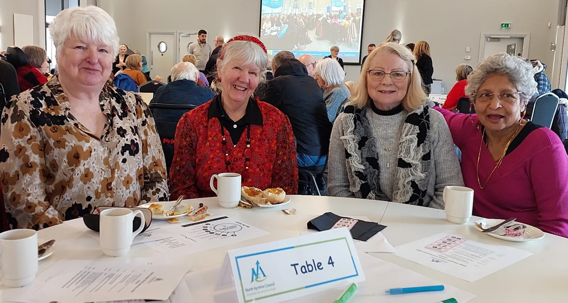Five older women sitting at a table at the Older People's Voice Conference with other table and older people in the background