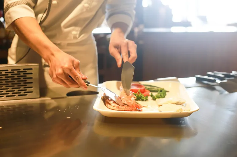 Chef preparing food dish with utensils on a plate on a stainless steel worktop.