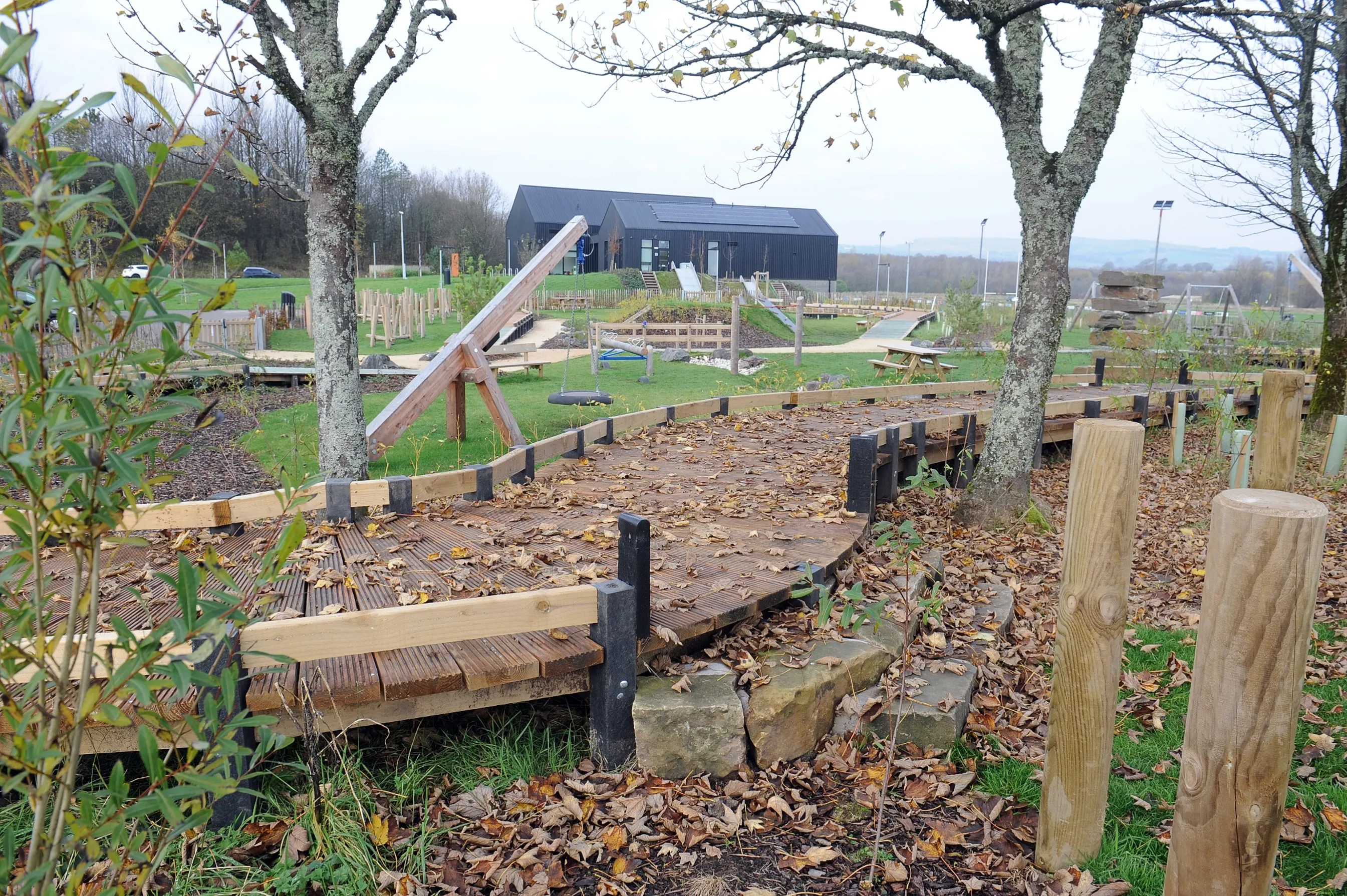 A raised wooden path with a covering of leaves, with a playpark and the Lochshore Park Hub in the background