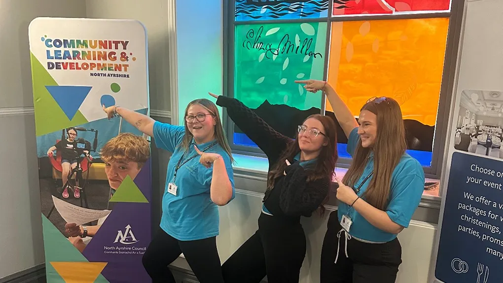 Three members of staff standing in front of a stained glass window pointing at a Community Learning and Development banner