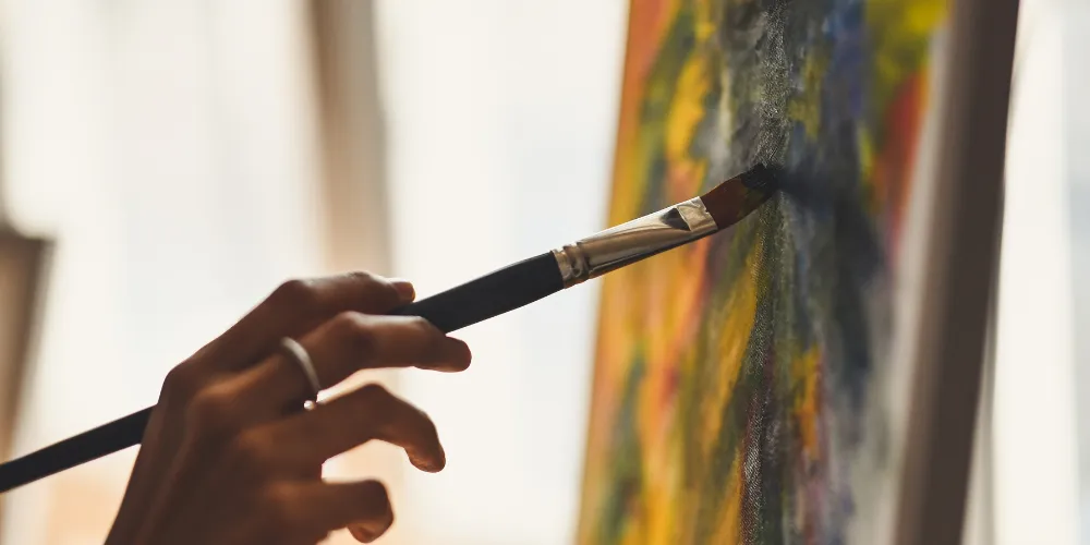 A paintbrush in an artist's hand against a painted canvas