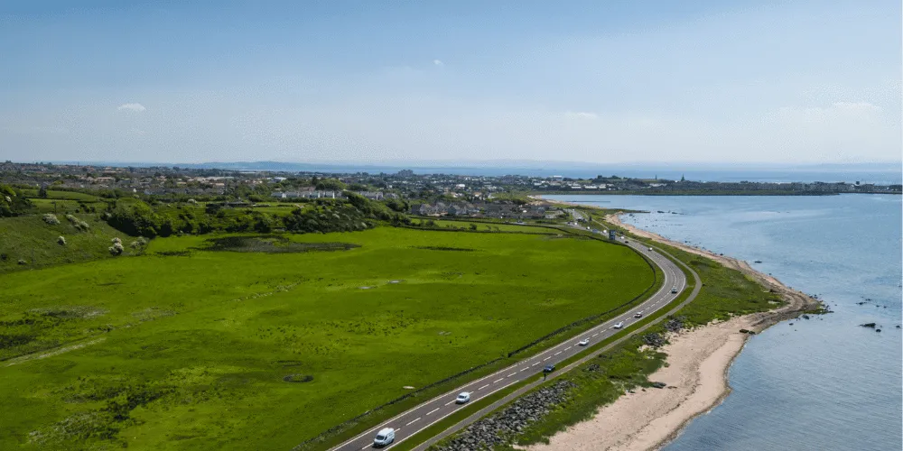 An aerial view of a coastal road with green fields and a town in the distance
