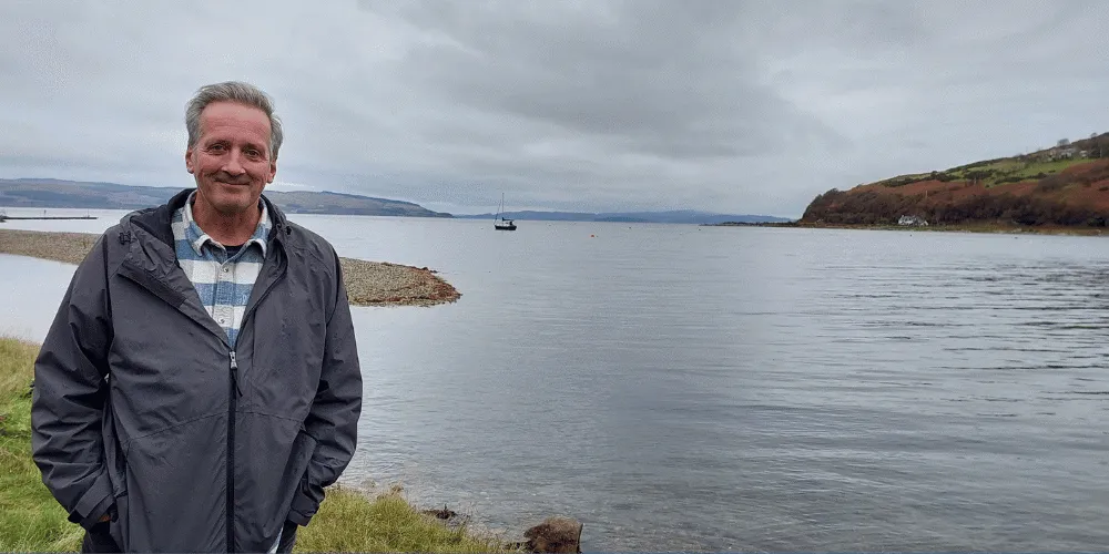 A man smiling at the camera with a boat bobbing about on water behind him