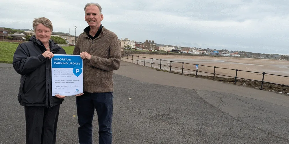 Two people holding a poster with the words 'important parking update' on a stretch of land next to a beach