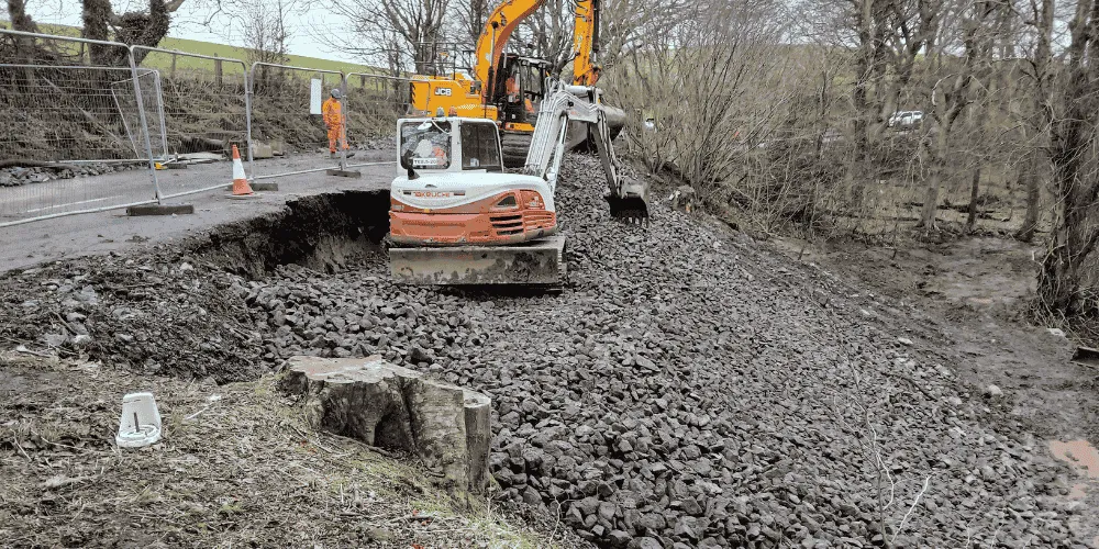 A piece of heavy digging machinery at the top of a slope next a road which has been coned off