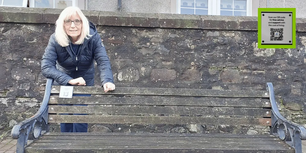 Councillor Marie Burns leaning on a street bench that has a QR code plaque attached to it.