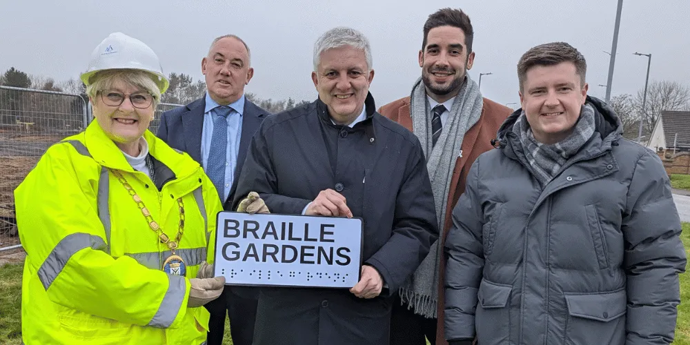 A group of five people, smiling, and holding a street sign which says Braille Gardens