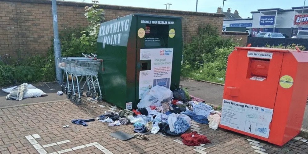 Two charity clothing banks, with shops in the background and burst bags of donations strewn in the foreground