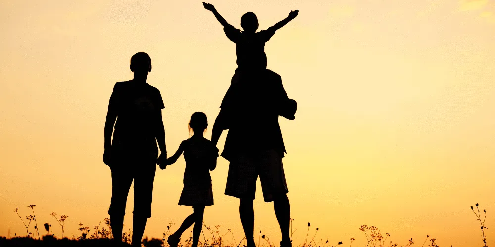 Silhouette of a family - two adults and two children - holding hands against an amber sky in the background
