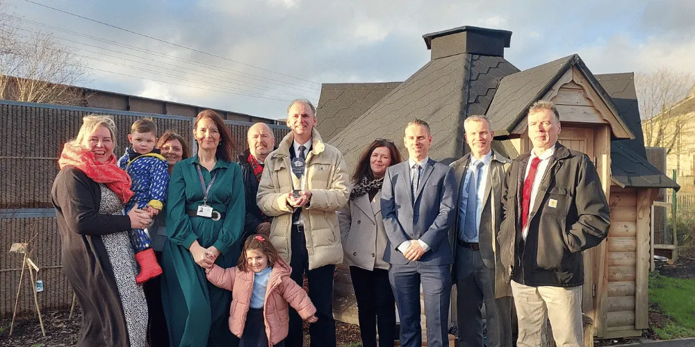 Men, women and children smiling while lined up in front of a wooden play house