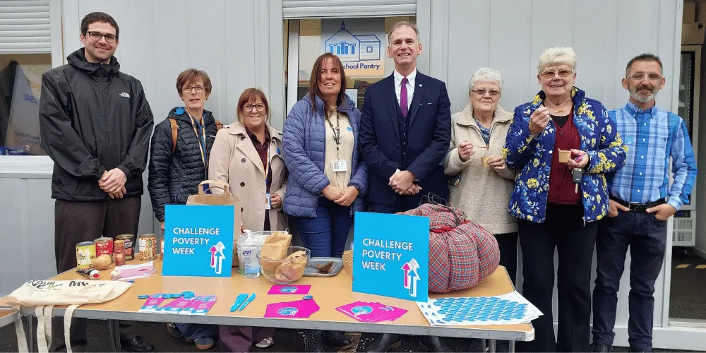 Eight people standing at a table with a pot of chilli on on it and Challenge Poverty Week materials