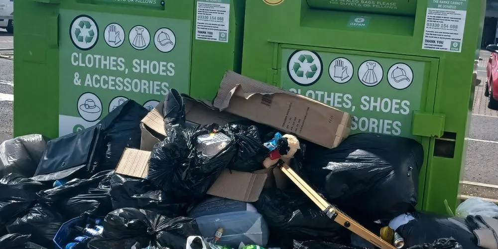Multiple black bin bags full of donations piled on the ground near two full clothing banks