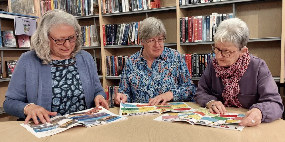 Three women sitting at a table looking at copies of a magazine, surrounded by books in a library 