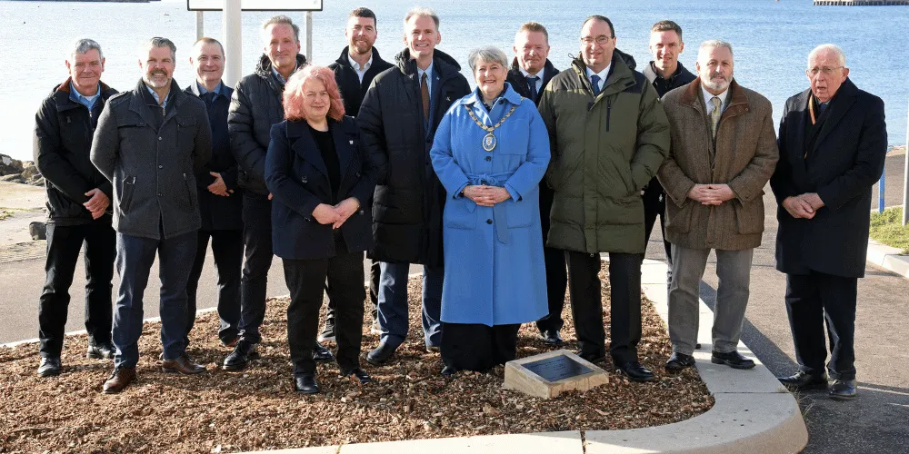 A group of men and women standing behind a small plaque, facing the camera, on a sunny day