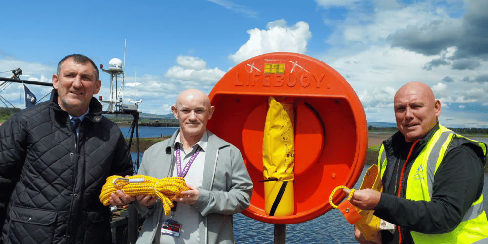 Three men standing in front of a life-saving buoy at a waterfront