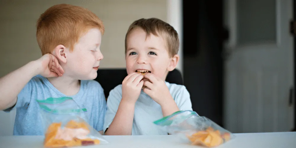 Two children sitting next to each other eating slices of orange from food bags
