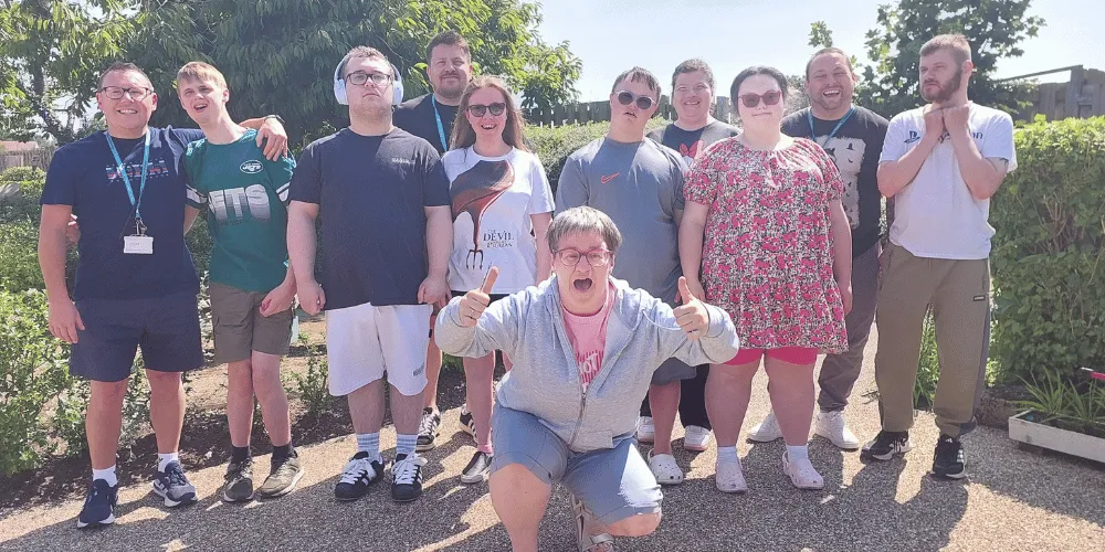 10 people standing in a row in a garden, with a woman kneeling in front making a "thumbs-up" sign.