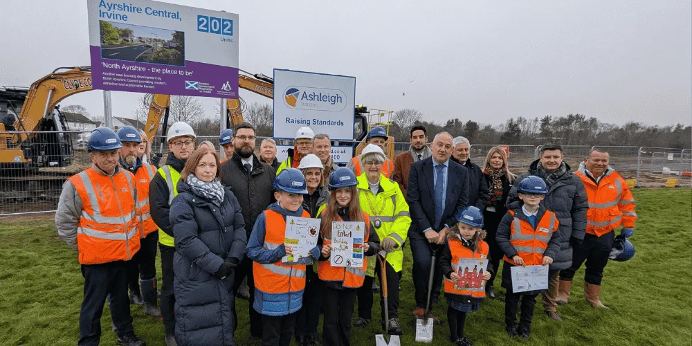 A large group of people stand on a grassed area in front of some signs showing new houses to be built