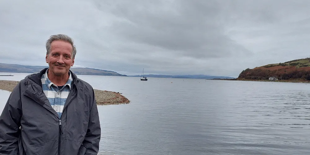 A man standing in front of a body of water with a boat bobbing on the water in the background