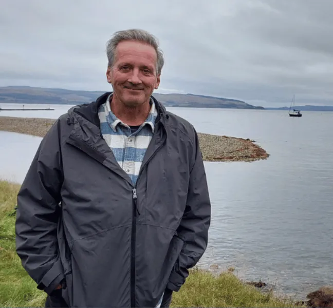A man smiling at the camera with a boat bobbing bout on water in the background
